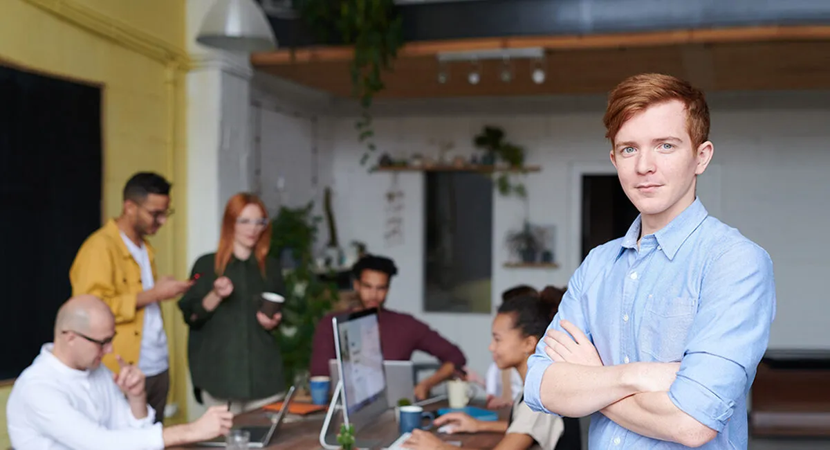 A man standing beside colleagues working at a table with laptops